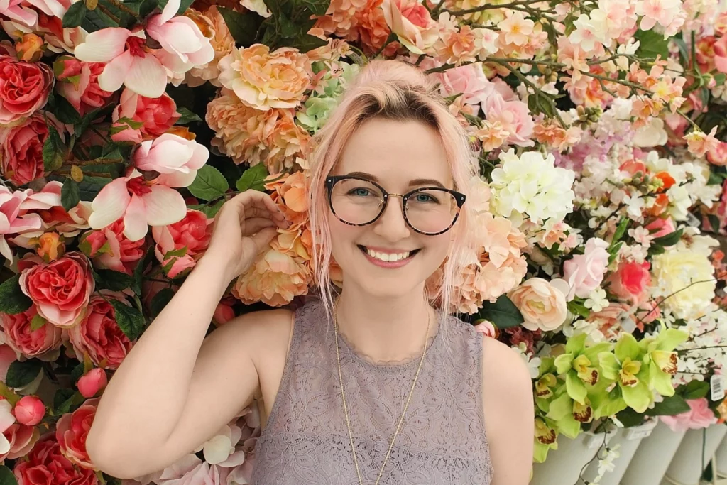 A smiling white woman with light pink hair and glasses standing in front of a wall of flowers