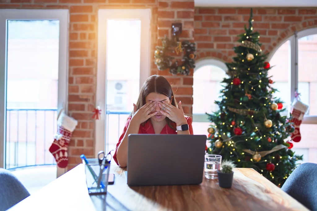 woman sitting at the table working with laptop at home around christmas tree rubbing eyes for fatigue and headache, sleepy and tired expression.